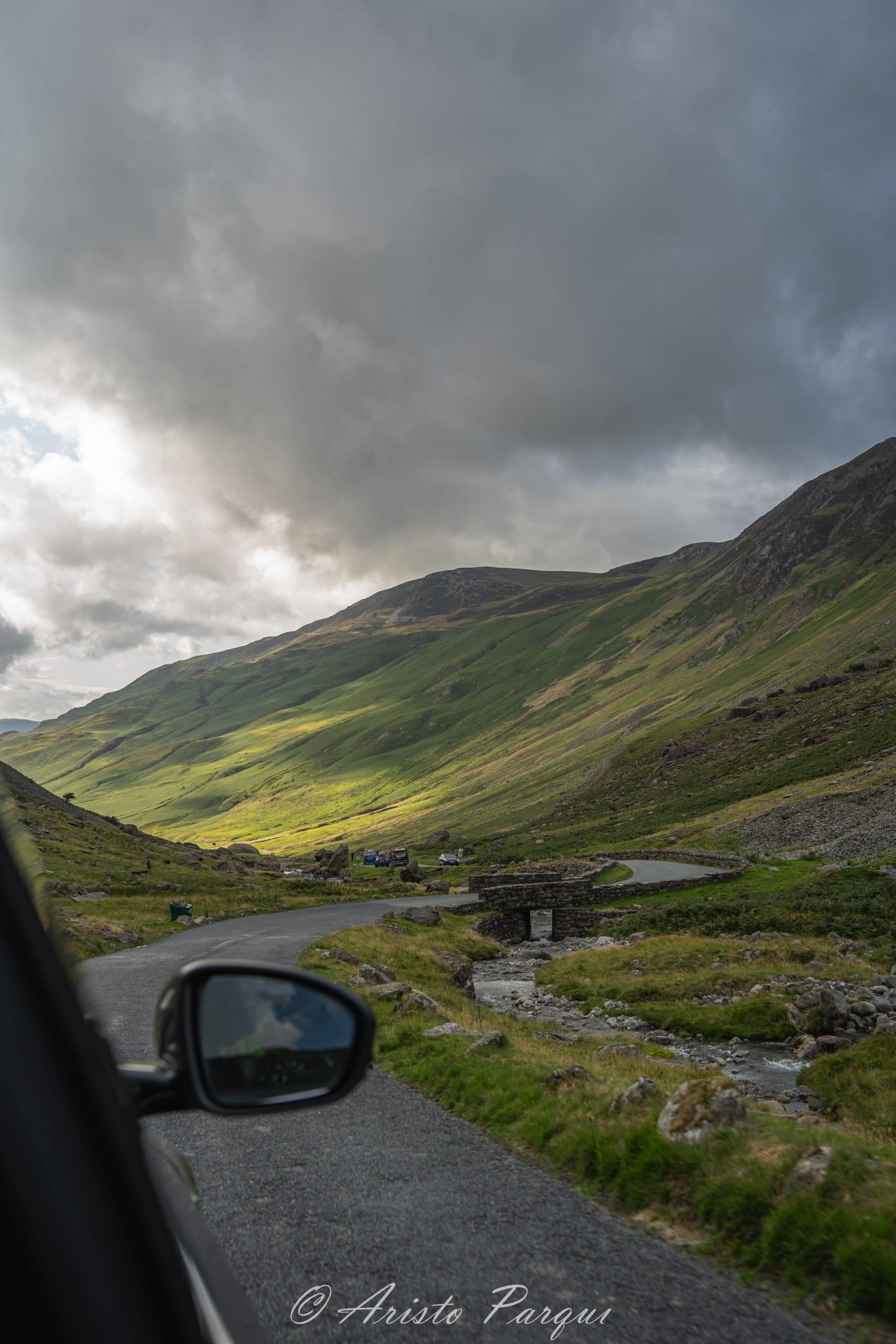 A stock photograph of a car driving on a road between green hills in Lake District, England.